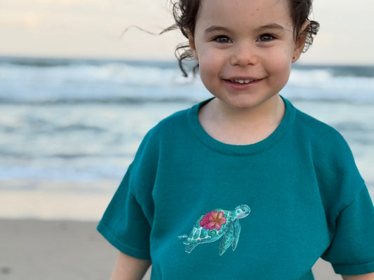 Child wearing a teal shirt with a turtle design on a beach. Eco-friendly toddler ocean tee inspired by Great Barrier Reef. limited edition