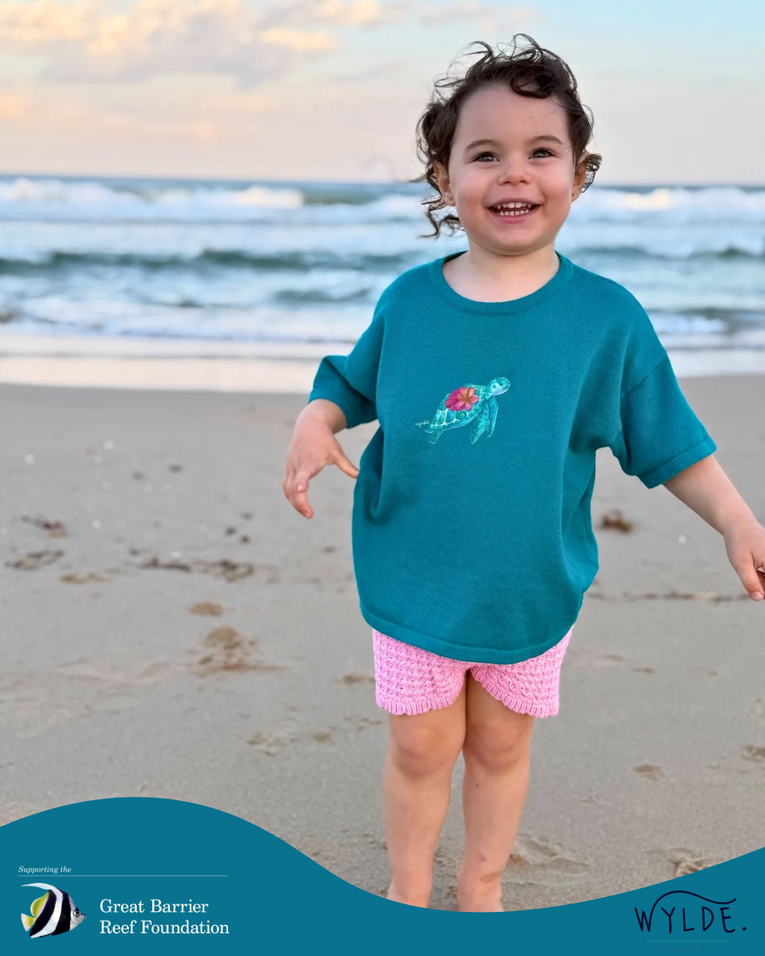 Child wearing a teal shirt with a turtle design on a beach, with 'Wylde' and 'Great Barrier Reef Foundation' logos.